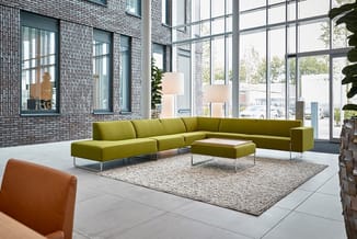 Groene modulaire hoekbank met ottomaan op beige vloerkleed in moderne kantoorlounge met grote ramen en bakstenen muur.