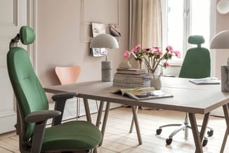 Modern kantoor met grijze tafel, groene bureaustoelen, witte hanglamp, roze bloemen in vaas en wandrek met foto's.