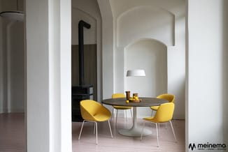 Ronde eettafel met donker blad en wit voetwerk, omgeven door vier gele stoelen met witte poten in minimalistisch interieur.
