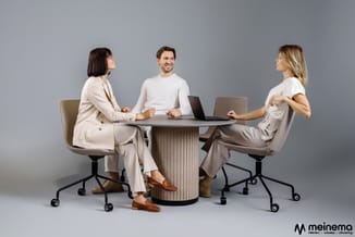 Drie personen zitten rond een ronde vergadertafel met verticale latten. Moderne draaistoelen in beige/taupe tinten. Laptops e
