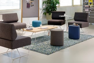 Moderne kantoorinrichting met grijze stoelen, blauwe en grijze poefjes, houten tafel, groen tapijt, plant en raampartijen.