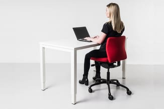 Vrouw zittend aan wit bureau met laptop, op rode en zwarte bureaustoel met wielen, tegen witte achtergrond.