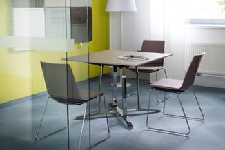 Moderne vergaderruimte met rechthoekige tafel, vier bruine stoelen met zadelzitting, glazen scheidingswand, gele accent muur