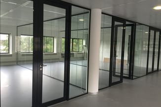 Glazen kantoorwanden met zwarte aluminium profielen in open kantoorruimte met witte vloer, witte plafond en natuurlijk licht.