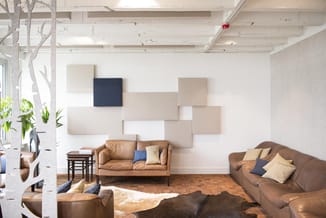 Kantoorlounge met bruine leren banken, akoestische wandpanelen in beige en wit, witte decoratieve boom, houten vloer en natuu