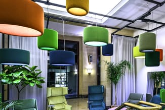 Moderne kantoorruimte met hanglampen in groen en oranje, gele en blauwe zitmeubelen, potplanten en gordijnen.
