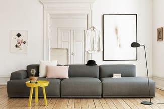 Grijze modulaire zitbank in ruim wit interieur met geel bijzettafeltje, zwarte vloerlamp en witte deuren op houten vloer.