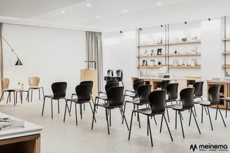 Moderne kantoorruimte met zwarte stoelen rond, houten kasten en wandplanken tegen witte muren, grijze vloer, hanglamp.