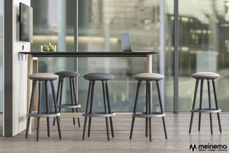 Zwarte bartafel met houten poten en vijf grijze barkrukken met kussen in modern kantoorinterieur met ramen.