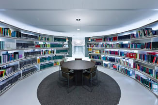 Ronde bibliotheekruimte met grijze tafel, twee donkergrijze stoelen, boeken in wit en grijs kastensysteem, hanglamp, LED-verl
