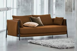 Bruine loungebank met donkergrijs metalen frame, sierkussens en grijze shagvloerkleed in modern kantoor met grote ramen.