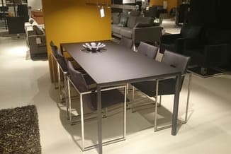 Moderne vergadertafel met grijs blad en metalen frame, omringd door grijze stoelen in showroom met gele wand.