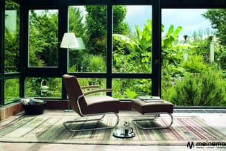 Modern loungegebied met bruinleren fauteuil, voetenbankje en glazen tafel op gestreept vloerkleed, grote ramen met groene tui