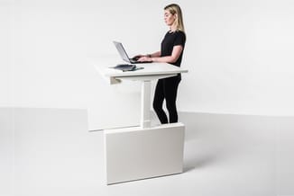 Vrouw werkend aan wit verstelbaar bureau met laptop in staande positie, geplaatst op wit onderstel in minimalistisch interieu