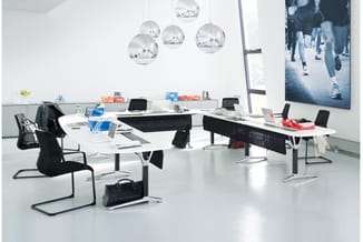 Modern kantoor met witte werkbladen, zwarte bureaustoelen, zilveren hangende bollampen, grote wandscherm en grijze vloer.