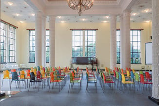 Gekleurde stapelstoelen in rood, oranje, geel, groen en blauw opgesteld in een klassieke zaal met kolommen, grote ramen en kr