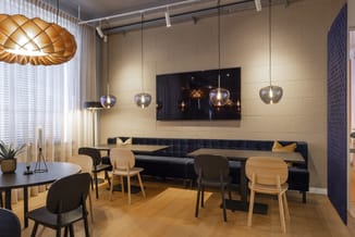 Modern lounge met zwarte hoekbank, houten stoelen aan ronde tafel, hangende glazen lampen, beige wandtegels en flatscreen TV.