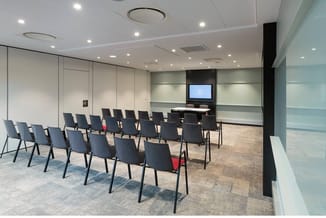 Moderne vergaderruimte met grijze stapelstoelen in rijen, whiteboard-wand, monitor aan zwarte kolom, spotlights en laminaatvl