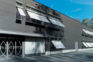 Modern grijs bakstenen kantoorgebouw met witte zonwering, glazen ingang met ronde raambeschermingen en helder daglicht.