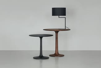 Twee ronde pedestal-tafels: één zwart metaal, één houtkleurig, met daarop een zwarte tafellampe met rechthoekige kap.