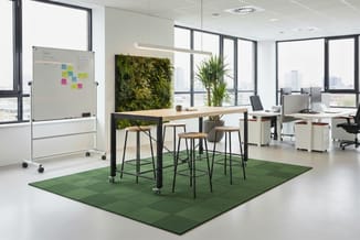 Moderne kantoorruimte met houten staantafel, zwarte barkrukken op groen tapijt, whiteboard met notities, groen muurbeplanting