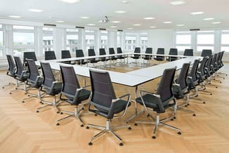 Moderne vergaderruimte met U-vormige witte vergadertafel en zwarte bureaisstoelen met wieltjes, grote ramen, houten vloer en