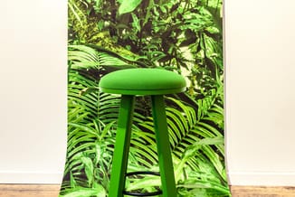 Groene barkruk met zacht zitoppervlak en drie poten, geplaatst voor een achtergrond met tropische plantenmotief.