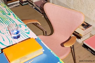 Roze gestoffeerde stoel met houten poten aan bureau met kleurrijke onderlegger, geel notitieboekje en glas water.