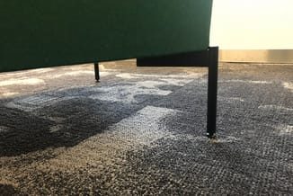Grijze en beige vloerbekleding in kantoorruimte met groene wand en zwarte palen, daglicht vanuit raam.