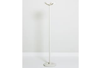 Staande vloerlamp met creme/beige kleurige metalen paal en voet, kleine lampenkop bovenaan, tegen witte muur.