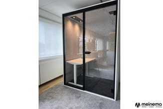 Moderne belcel met zwart aluminium frame en donker getint glas. Binnen een houten tafel en witte poten. Grijze vloerbekleding