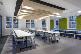 Moderne vergaderzaal met lange witte tafel, blauwe stoelen, beige plafondlampen, groene muswanden en grote ramen.