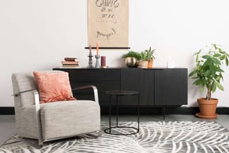 Grijze fauteuil met roze kussen op geometrisch vloerkleed, zwart dressoir met planten, kleine tafel en wanddecoratie in moder