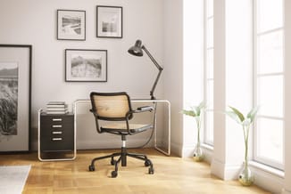 Modern kantoor met zwarte bureaustoel met rattan rugleuning, wit bureau, zwart kastje, bureaulamp en potplanten bij raam.