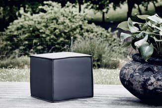 Zwarte kubusvormige poef op houten terras, omgeven door tuinplanten en een zwarte bloempot met groen blad.