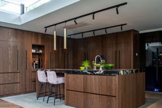 Moderne keuken met donkerbruin houten kasten, zwart werkblad, eiland met twee grijze barkrukken, gouden hanglampjes en skylig