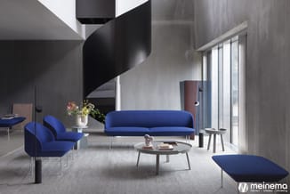 Moderne lounge met blauwe zitmeubelen, ronde tafel, spiraalvormige trap, grijze betonnen wanden en grote ramen.