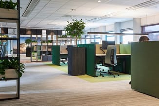 Modern kantoor met groene akoestische schermen, werkplekken, houten vloer, plantenaccenten en natuurlijk licht.