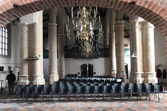 Zwarte stoelen in rijen opgesteld in een kerkinterieur met bakstenen zuilen, kroonluchter en bakstenen gewelfd plafond.