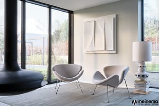 Moderne loungeruimte met twee grijze fauteuils, zwarte kolom, grote ramen, wit kunstwerk aan muur en witte tafellamp met stap