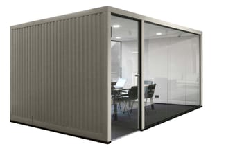 Grijze kantoorbelcel met golfplaat zijwanden, glazen deur en voorzijde. Binnenin bureau met laptop, twee stoelen en verlichti