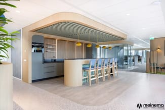 Modern kantoorkeukentje met licht houten kastenwand, zwarte keukenapparatuur, gouden hanglampjes boven een bartafel met blauw