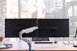 Ergonomische monitorarm in zilver voor dubbele monitoren op houten bureau, met toetsenbord, muis en notitieblok.