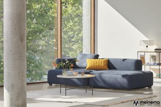 Moderne woonkamer met donkerblauwe hoekbank, geel kussen, ronde salontafel, betonnen zuil, grote ramen met uitzicht op groen,