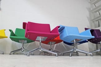 Rij gekleurde kantoorstoeien in groen, roze, rood, blauw en paars met zilveren draaipoten tegen witte muur.