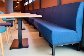 Blauwe zitbank met hoge rugleuning in moderne kantooromgeving, wit tafelblad op zwart onderstel, houten stoelen, oranje muren