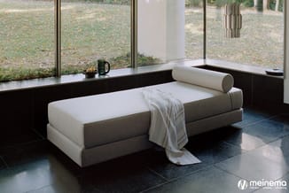 Grijsbeige loungebank met witte kussens in minimalistische ruimte met grote ramen en zwarte tegelvloer.