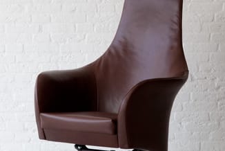 Bruine leren draaifauteuil met hoge rugleuning op vierpotig metalen onderstel, tegen witte bakstenen muur.