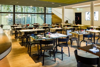 Modern restaurant met zwarte stoelen, witte en grijze tafels, houten vloer, grote ramen en gele trapopgang.