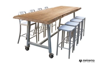 Hoge tafel met natuurlijk houten blad en metalen onderstel op wielen, omgeven door vier barkrukken met grijze zitting.